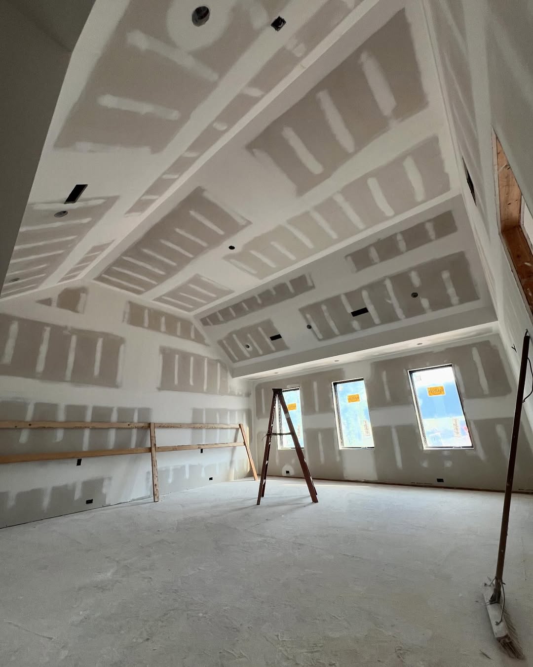 A large room with a high, vaulted ceiling in the midst of drywall installation, with a wooden ladder placed near unfinished walls.