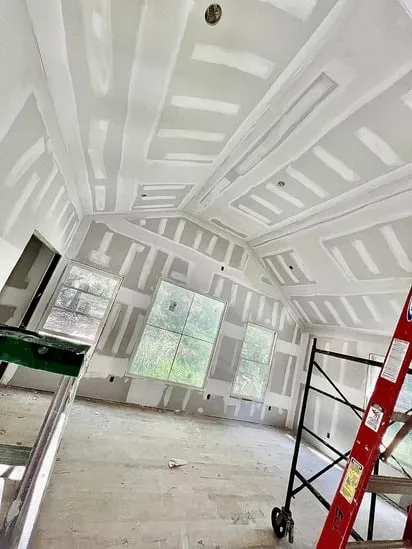 An interior room view showing a vaulted ceiling and walls with fresh joint compound applied over all seams, part of the drywall taping process. Scaffolding, a red ladder, and large windows are visible.