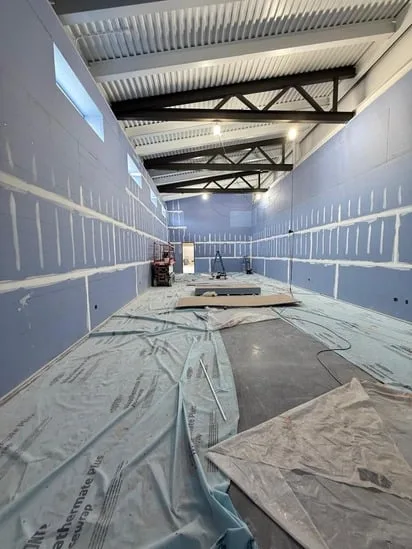 Commercial drywall finishing underway in a spacious room, showing freshly mudded seams and protective floor covering.