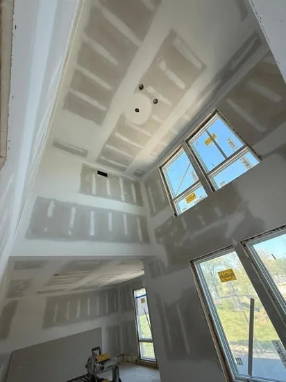 Drywall taping and mudding completed on a high vaulted ceiling and walls surrounding large modern windows.